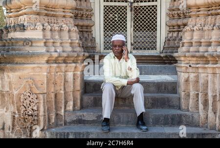 Junagadh, Gujarat, India - December 2018: An elderly Indian man wearing a skullcap talks on the phone while sitting on the steps of an ancient monumen Stock Photo
