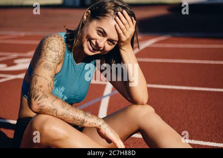 Cheerful wet athlete girl with tattooed hand in sportswear joyfully looking in camera while resting after running on city stadium Stock Photo