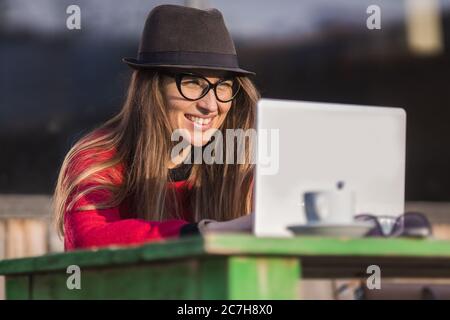young woman with black glasses and hat working on laptop at outdoor coffee shop table Stock Photo
