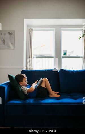 Preschool age boy laying on blue couch using tablet to learn Stock Photo