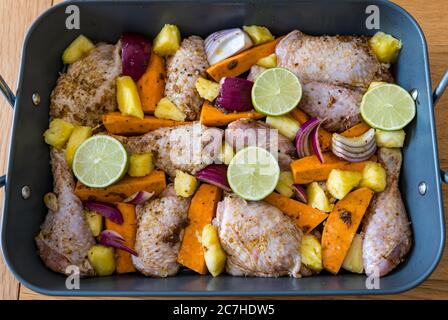 Jerk chicken thighs & drumsticks tray bake in roasting tin ready to cook with vegetables: sweet potatoes, red onions, lime & pineapple chunks Stock Photo