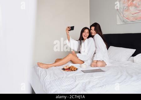 Two smiling women in white bathrobes happily taking photos together on cellphone with delicious pastry and coffee. Attractive girls having breakfast Stock Photo