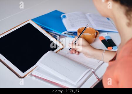 Close up photo of girl sitting at the table with tablet and cup of coffee writing in notebook while studying at home Stock Photo