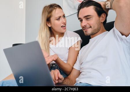 Young blond woman amazedly looking at smiling brunette man happily using laptop. Young couple in white T-shirts sitting on bed together at modern home Stock Photo