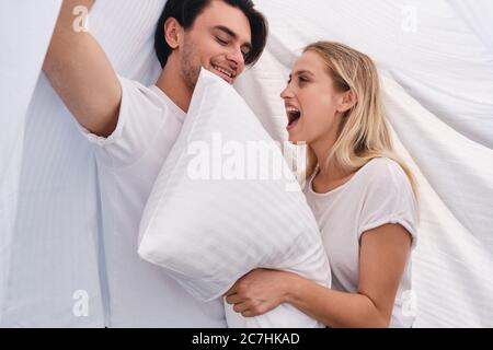 Handsome brunette man and pretty cheerful blond woman with white pillow in hands happily sitting and covering with blanket in bed at cozy home Stock Photo