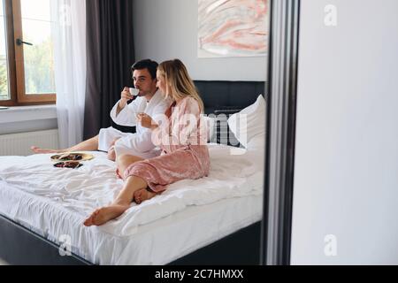 Handsome brunette man in white bathrobe thougtfully looking aside while drinking coffee with pretty blond woman in pink velevt robe. Young couple Stock Photo
