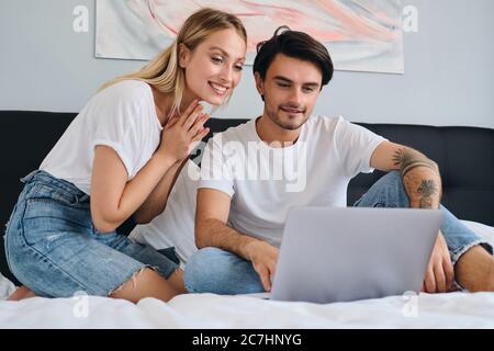 Pretty blond woman and attractive brunette man happily working on laptop together. Young beautiful couple in white T-shirts sitting on bed at home Stock Photo