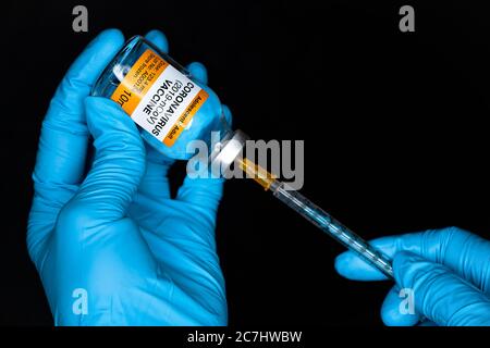 Hand in blue gloves holding hypodermic syringe and coronavirus vaccine (sticker own design, not real product), injecting it closeup detail, black back Stock Photo