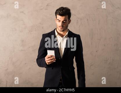 A serious handsome young businessman in a black suit using a smartphone in front of a grey wall in a studio. Stock Photo