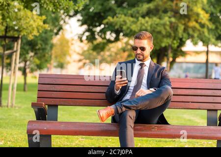 A handsome young businessman sitting on a bench and using his phone on a sunny day. Stock Photo