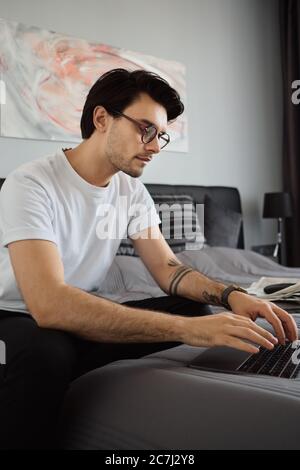 Young attractive pensive brunette man in T-shirt and eyeglasses sitting on bed and dreamily working on laptop at modern home Stock Photo