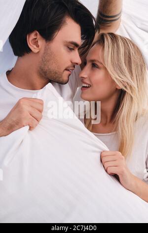 Young brunette man and beautiful blond woman dreamily looking at each other with pillow in hands lying under blanket in bed at home Stock Photo