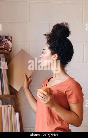 Young pretty woman with dark curly hair in T-shirt holding cup of coffee in hand thoughtfully choosing book from bookshelf at cozy home Stock Photo