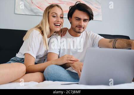 Pretty cheerful blond woman and attractive brunette man happily watching movie on laptop together. Young cute couple in white T-shirts sitting on bed Stock Photo