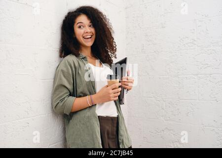 Young cheerful woman with dark curly hair in khaki shirt holding laptop and cup of coffee to go in hands and joyfully looking in camera isolated Stock Photo
