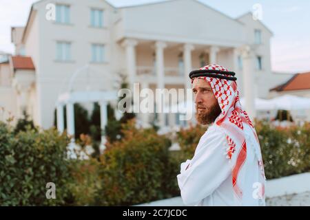 Handsome middle eastern arab muslim man in traditional clothing standing in front of the white beautiful modern house Stock Photo