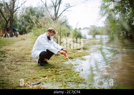 Male caucasian scientist biologist and researcher in protective suit with mask taking water samples from polluted river. Stock Photo