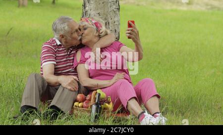 Family weekend picnic. Old people, merry senior caucasian couple using smartphone makes selfie. Remote online video chat call on phone. Elderly man, woman. Husband and wife in park. Grandparents Stock Photo
