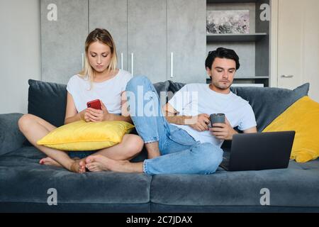 Young brunette man holding cup in hand thoughtfully looking in laptop while pensive blond woman near using cellphone. Attractive couple sitting on Stock Photo