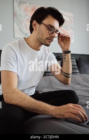 Young handsome brunette man in T-shirt and eyeglasses sitting on bed thoughtfully working on laptop at modern home alone Stock Photo