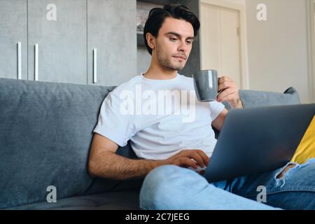 Young attractive brunette man in white T-shirt holding cup in hand thoughtfully working on laptop sitting on sofa at modern cozy home Stock Photo