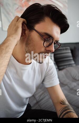 portrait of young handsome brunette man in T-shirt and eyeglasses sitting on bed at modern home and thoughtfully looking aside Stock Photo