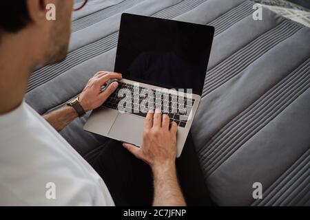 Close up photo of man sitting on bed and working on modern laptop at home Stock Photo