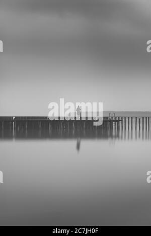 Vertical greyscale shot of a lonely person standing on a wooden dock near the sea Stock Photo