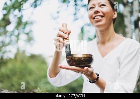 Young smiling woman playing on the brass Tibetan singing bowl outdoor. Sound therapy and meditation Stock Photo