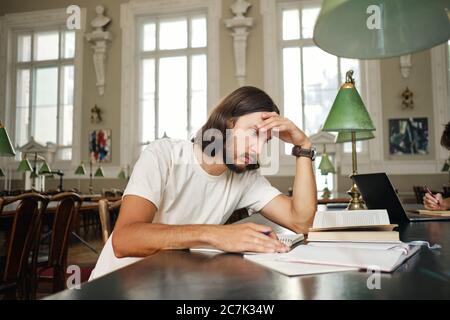 Young tired male student thoughtfully preparing to exam while studying in library of university Stock Photo