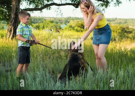 Young cheerful family is walking with his dog a German shepherd in the meadow. Mom and son spend time together and have fun in nature with a pet. Stock Photo