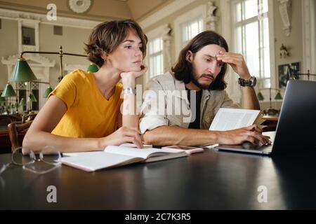 Two young pensive students thoughtfully working on study project with laptop and books in library of university Stock Photo