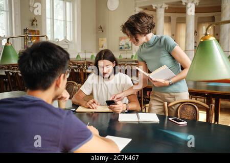 Young female tutor with book helping casual students with study during lesson in library of university Stock Photo