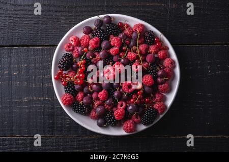 Tasty fresh ripe raspberry, blackberry, gooseberry and red currant berries in plate, healthy food fruit on dark wooden background, top view Stock Photo