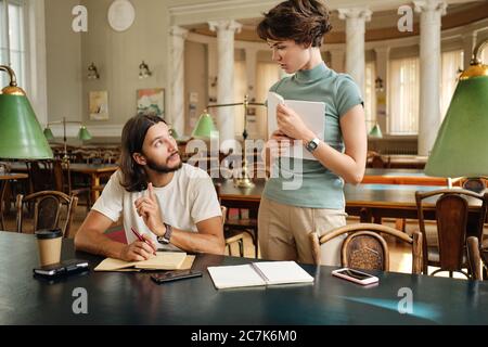 Attractive tutor with book thoughtfully discussing with young student new theme during lesson in library of university Stock Photo