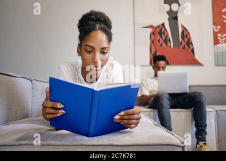 Beautiful African American girl lying on sofa dreamily reading book while boyfriend on background working on laptop at home Stock Photo