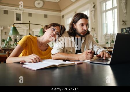 Two young students tiredly working on study project with laptop in library of university Stock Photo