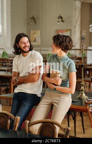 Young positive casual students with notepad leaning on table while happily talking during study break in library of university Stock Photo
