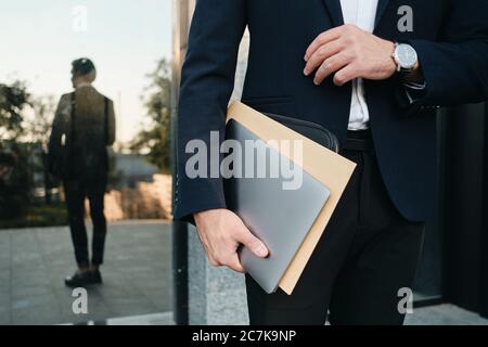 Close up man in classic suit and wristwatch holding laptop and big envelope in hand with reflection on background Stock Photo