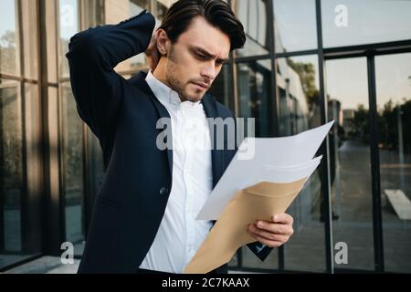 Young serious bearded brunette man in white shirt and classic jacket holding hand near head standing outdoor and thoughtfully reading documents with Stock Photo