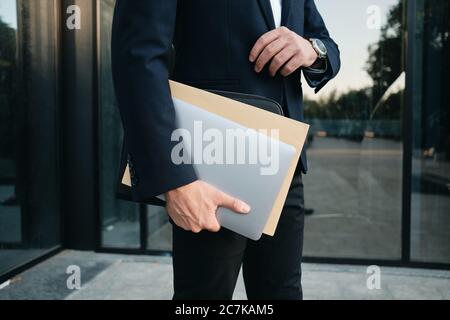Close up man in classic black suit standing outdoor alone holding laptop and big envelope in hand Stock Photo