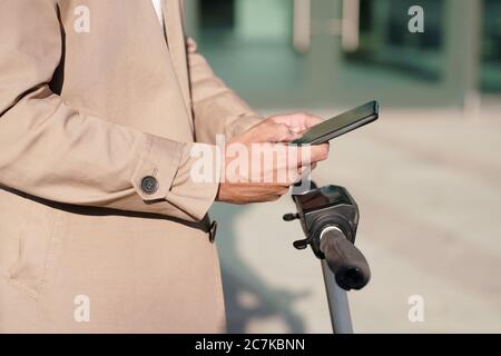 Hands of young mobile businessman in beige trenchcoat messaging in smartphone over handles of electric scooter in urban environment Stock Photo