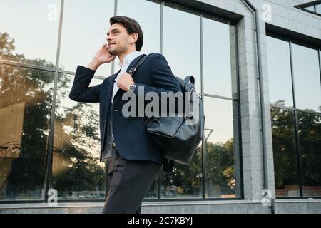 Young beautiful smiling bearded brunette man in white shirt and classic suit with wireless earphones and backpack on shoulder dreamily walking outdoor Stock Photo