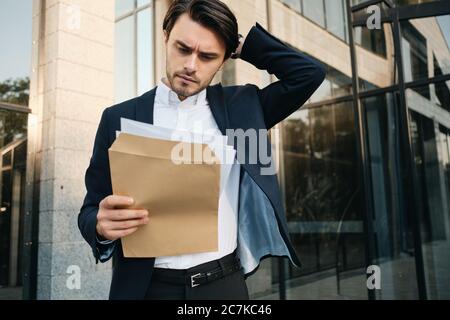 Young serious bearded brunette man in shirt and classic jacket thoughtfully looking on envelope with documents and glass building on background Stock Photo
