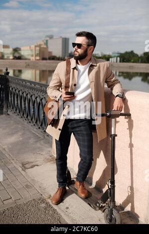 Young man in elegant stylish casualwear and sunglasses standing on bridge by riverside with urban environment on background Stock Photo