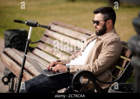 Young businessman with mobile gadget surfing in the net while sitting on wooden bench in park on warm day with his scooter near by Stock Photo