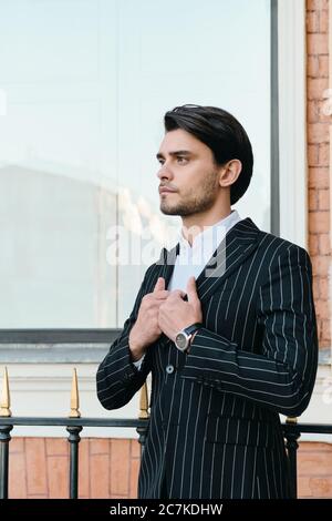 Young attractive bearded brunette man in white shirt and classic striped jacket thoughtfully looking aside standing on city street Stock Photo