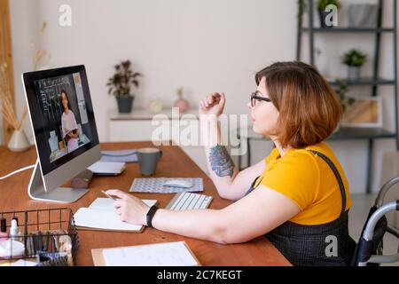 Young disable teacher or educational consultant in wheelchair sitting by desk in front of computer and talking to mixed-race schoolgirl Stock Photo