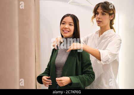 Young fashion consultant confidently helping trying on cardigan to pretty Asian girl in store