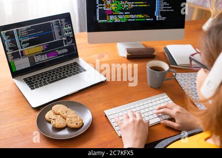 Young female programmer sitting by workplace at home, having tea with cookies and typing on computer keypad while analyzing data Stock Photo